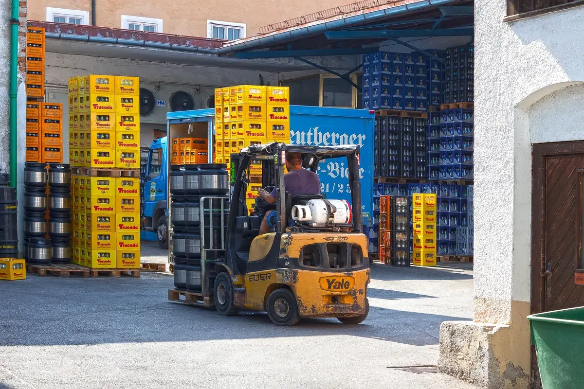 A man operates a forklift, moving black barrels in an outdoor warehouse. Stacks of colorful crates and a blue delivery truck surround the scene, conveying a busy work atmosphere.