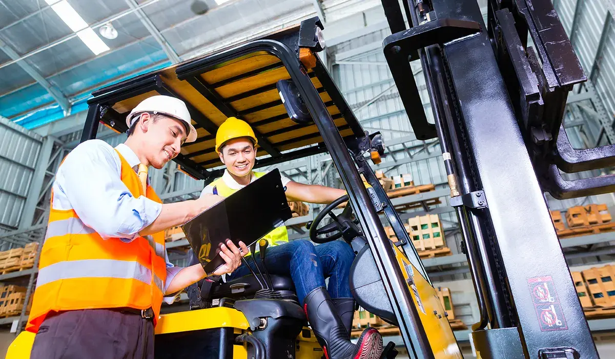Two warehouse workers in hard hats and safety vests discuss a clipboard near a forklift. One sits in the forklift, both appear engaged and smiling.
