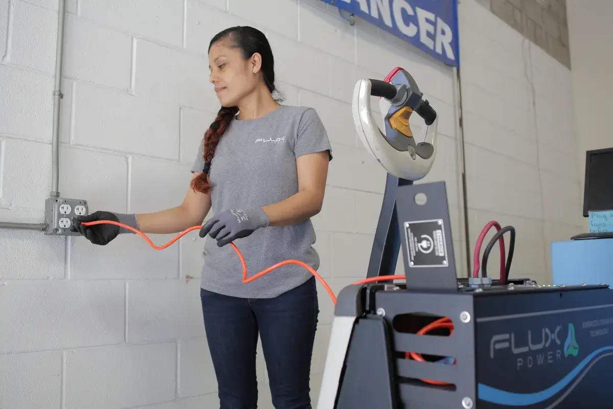 A woman in a gray "Flux" shirt and gloves plugs an orange cable into a wall socket beside a charging station. She appears focused and calm.